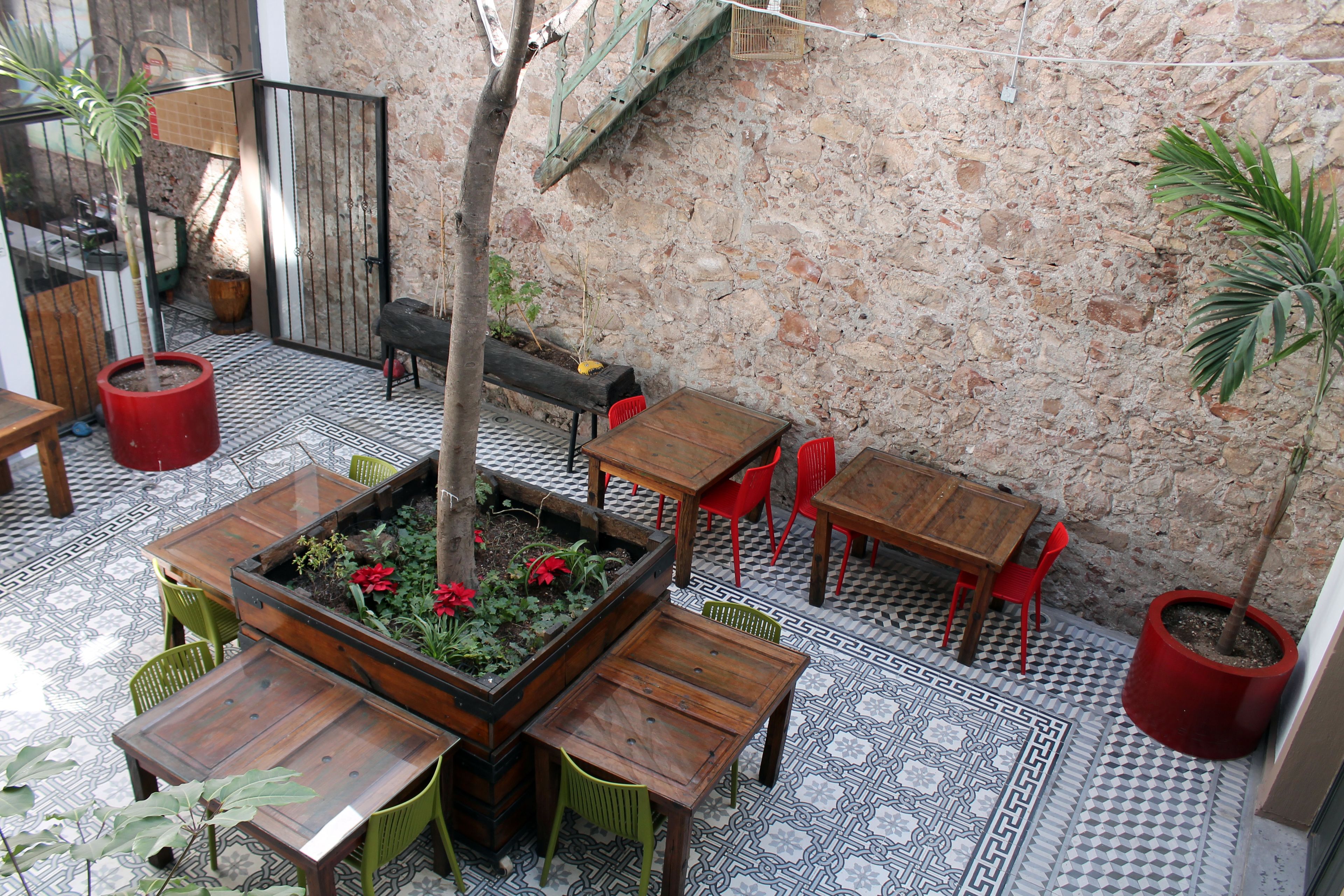 Un'area patio esterna con tavoli e sedie di legno, un piccolo aiuola con piante e un muro in pietra sullo sfondo.