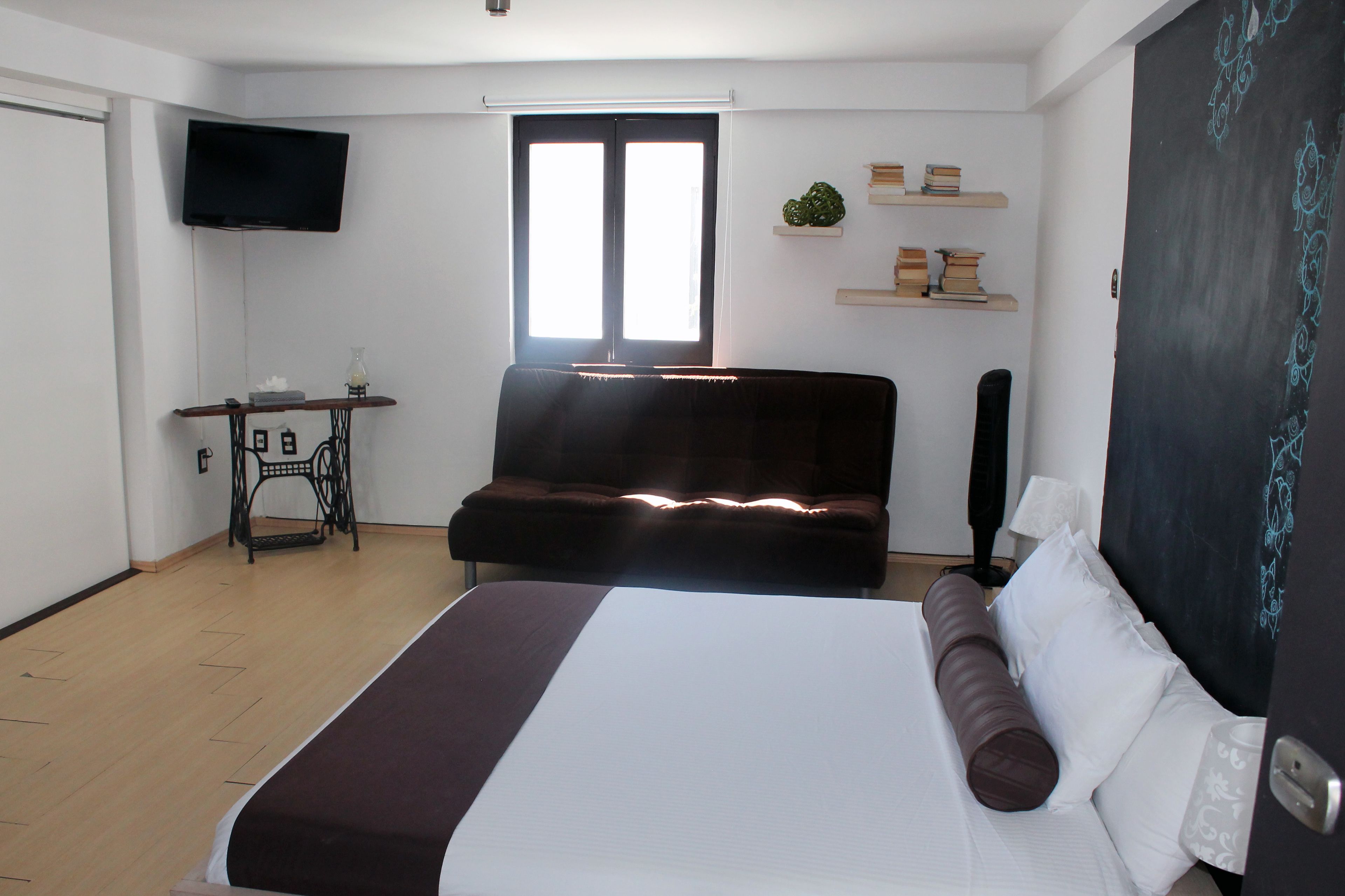 A hotel room with a bed, a sofa, a TV mounted on the wall, a window with blinds, and a chalkboard with writing.