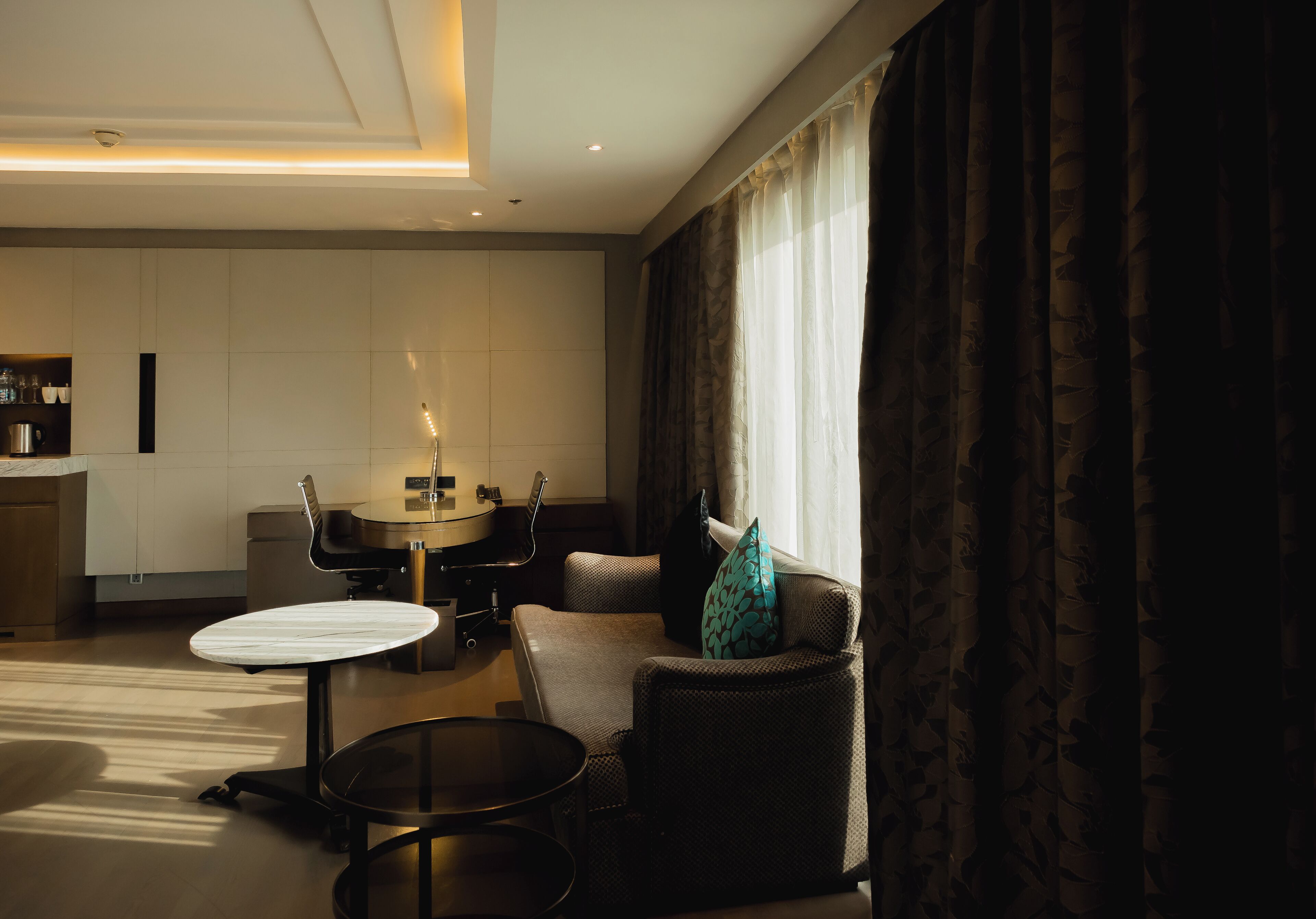 A modern hotel room with a sofa, armchair, round coffee table, and a dining table with chairs.