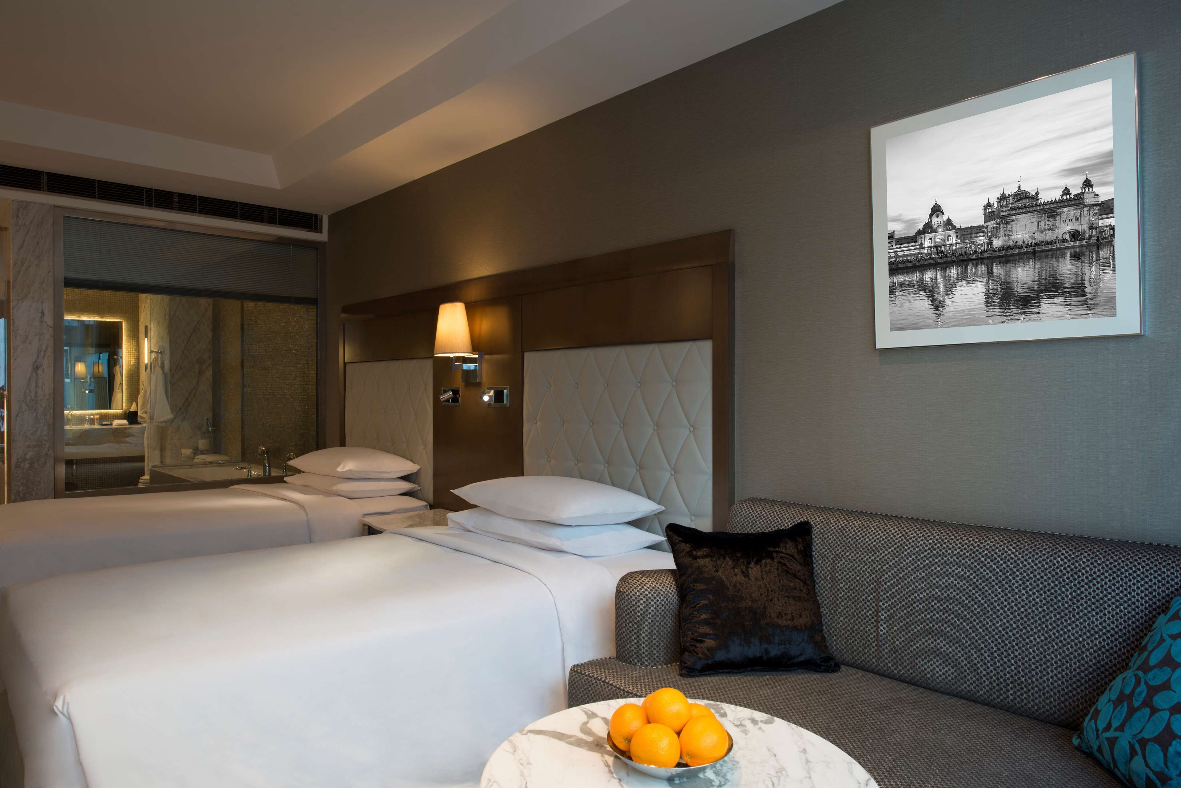 A hotel room with a large bed, a smaller bed, a sofa, a round table with a bowl of fruit, a wall-mounted lamp, and a framed picture on the wall.