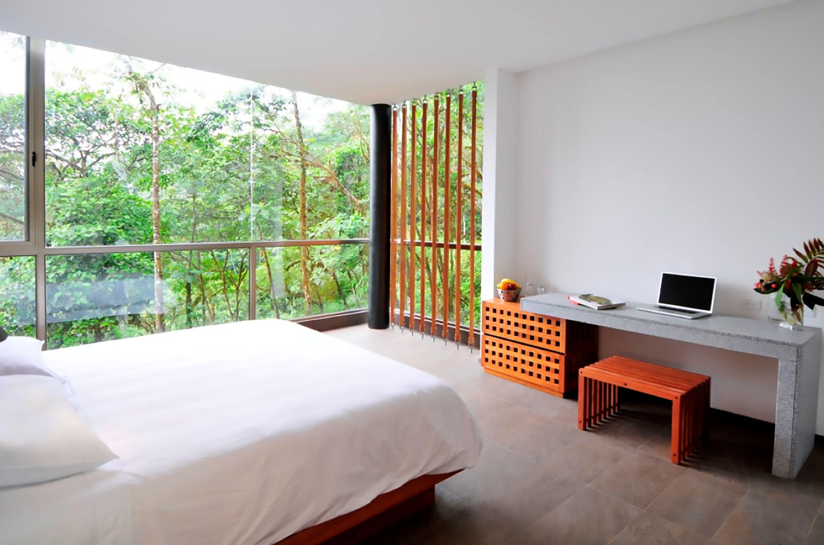 A bedroom with a large bed, a desk with a laptop, and a view of lush greenery outside.