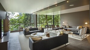 A spacious living area with a modern design, featuring a grey sofa, armchairs, a coffee table, and a dining table. Large windows provide a view of lush greenery outside.