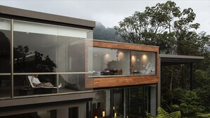 A modern glass and wood structure with a person relaxing on a balcony, surrounded by lush greenery and a mountainous backdrop. - Mashpi Lodge (Pacto)