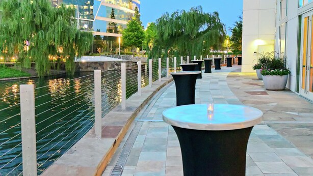 A riverside walkway with outdoor seating, a water feature, and modern buildings in the background. - Hilton Dallas/Plano Granite Park (Plano)