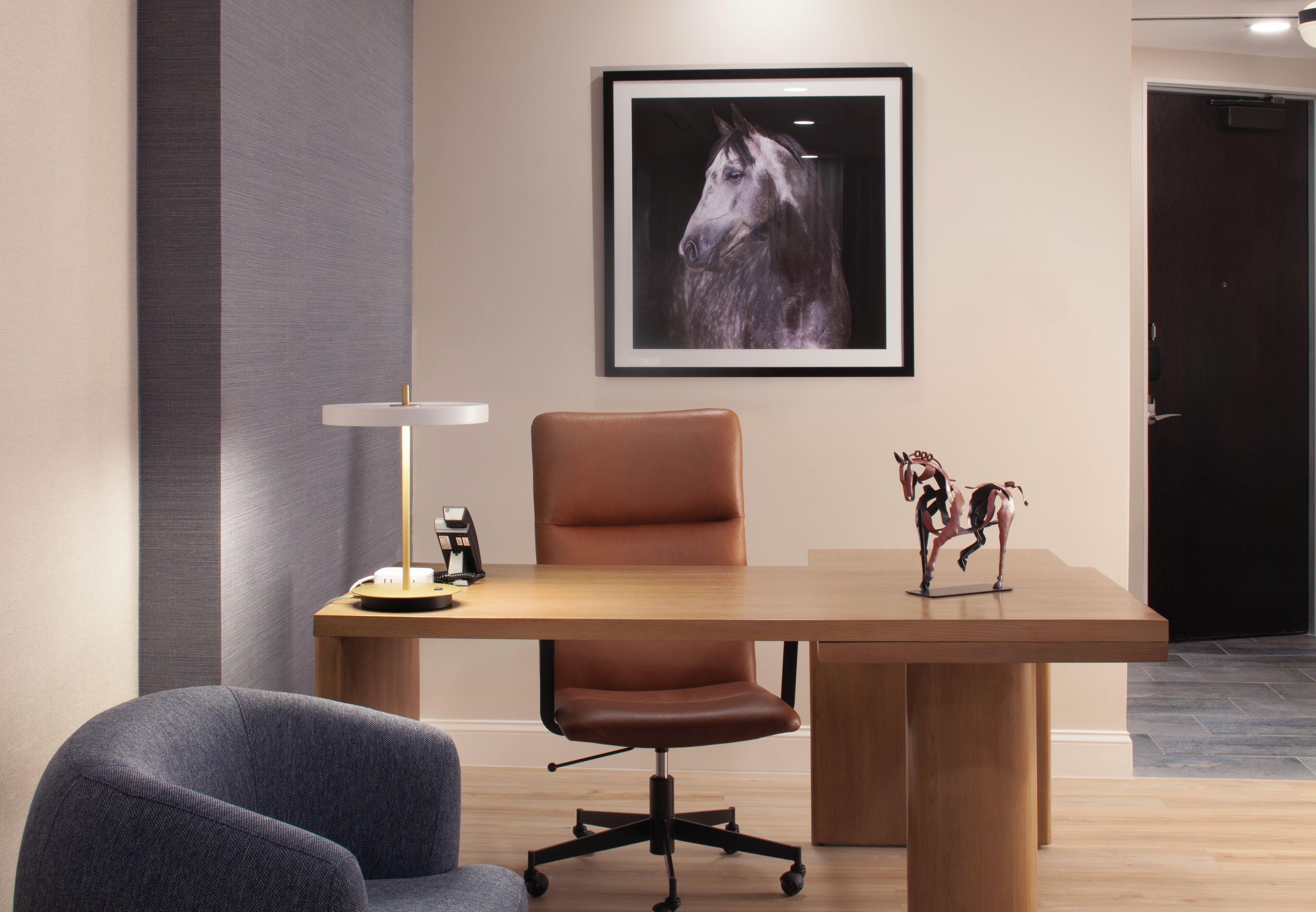 A conference room with a wooden table, a brown leather chair, a grey armchair, a framed horse portrait, and a small horse figurine.