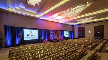 Un salón de conferencias con filas de sillas, un escenario con un logotipo de Hilton de fondo y una lámpara de araña moderna.