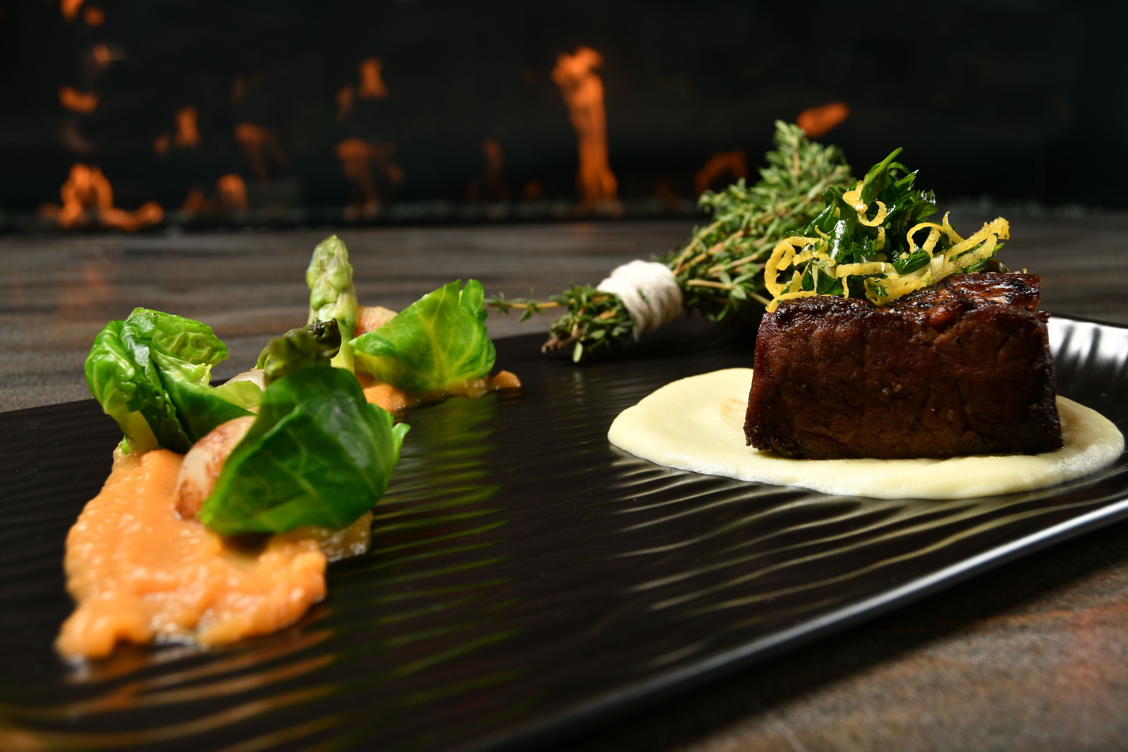 A black plate with a piece of meat, greens, and a sauce.