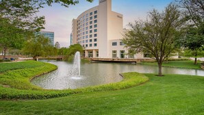 A hotel with a fountain in the courtyard, surrounded by greenery and a body of water. - Hilton Dallas/Plano Granite Park (Plano)