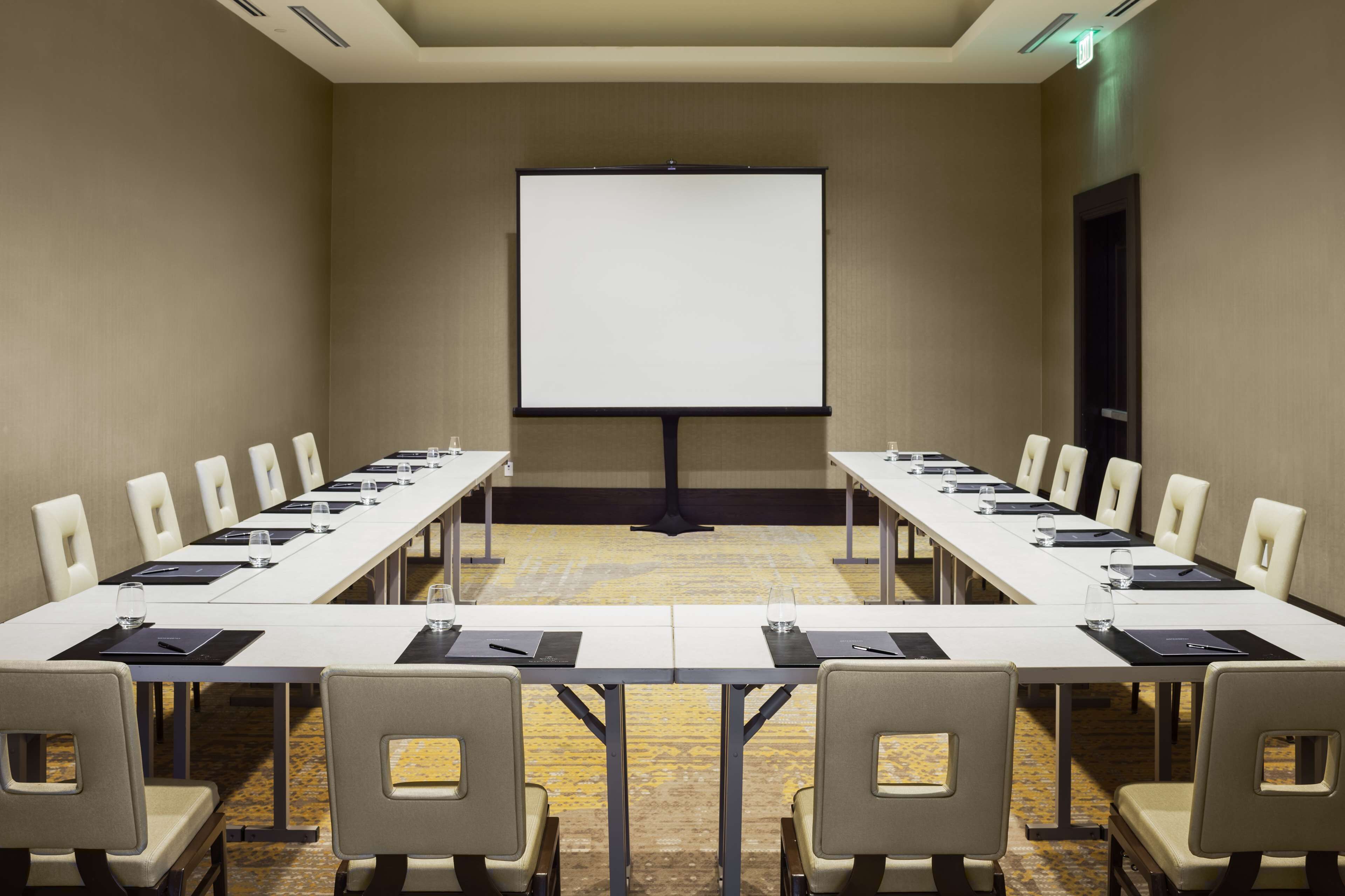Un salón de conferencias con una larga mesa, sillas, una pantalla de proyector y piso alfombrado.