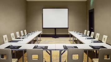 Un salón de conferencias con una larga mesa, sillas, una pantalla de proyector y piso alfombrado.