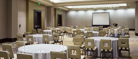 A conference room with multiple round tables and chairs, a projector screen, and a carpeted floor.