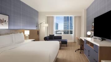 Habitación de hotel con una cama grande, un escritorio, una silla, un televisor y una ventana con vista a la ciudad.