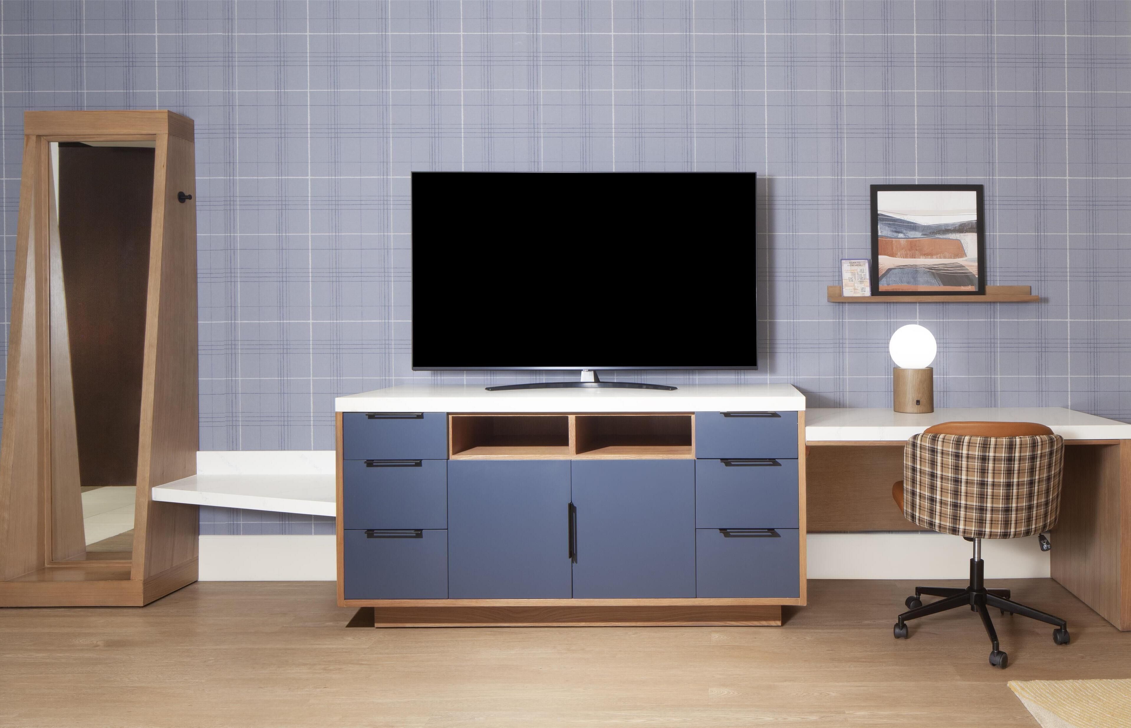 A modern bedroom with a wooden wardrobe, a flat-screen TV, a desk with a lamp, and a chair.