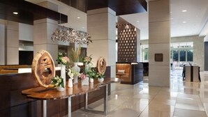 A modern reception area with a wooden reception desk, a decorative chandelier, and a seating area with a patterned wall. - Hilton Dallas/Plano Granite Park (Plano)