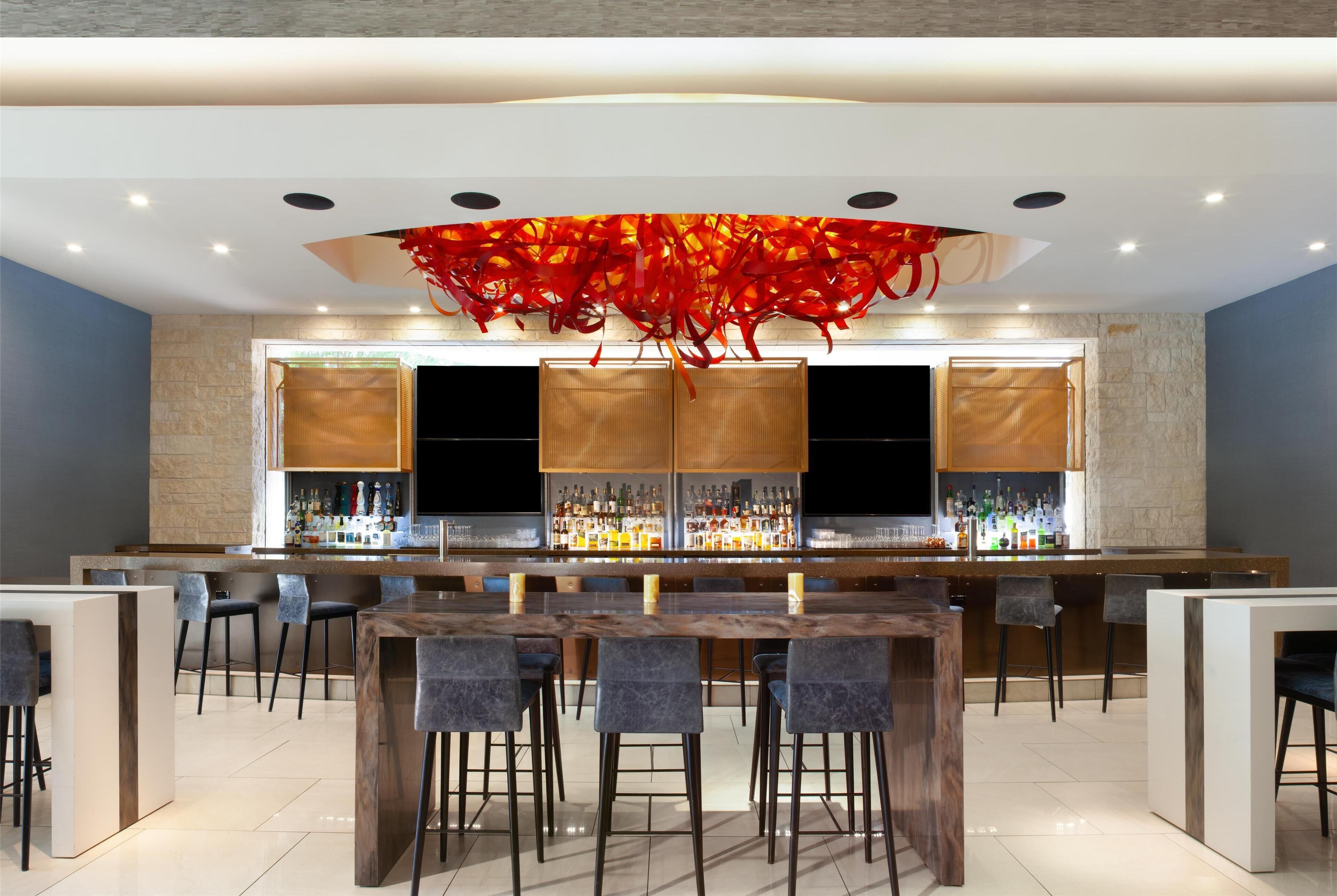 A modern bar area with a long counter, multiple bar stools, and a decorative red chandelier.