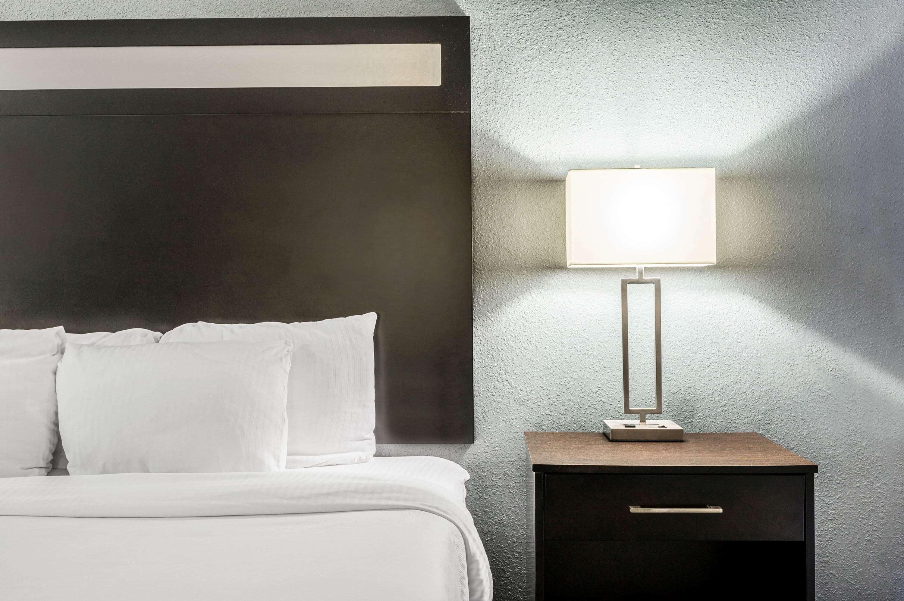 Een netjes opgemaakt bed met witte lakens en kussens, een donker hoofdbord en een nachtkastje met een lamp en een klein doosje.