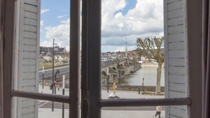Una vista attraverso una finestra con persiane, che mostra un ponte in pietra su un fiume, edifici e un cielo sereno.