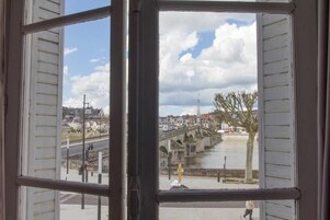 Une vue à travers une fenêtre à volets, avec un pont de pierre enjambant une rivière, des bâtiments et un ciel dégagé.