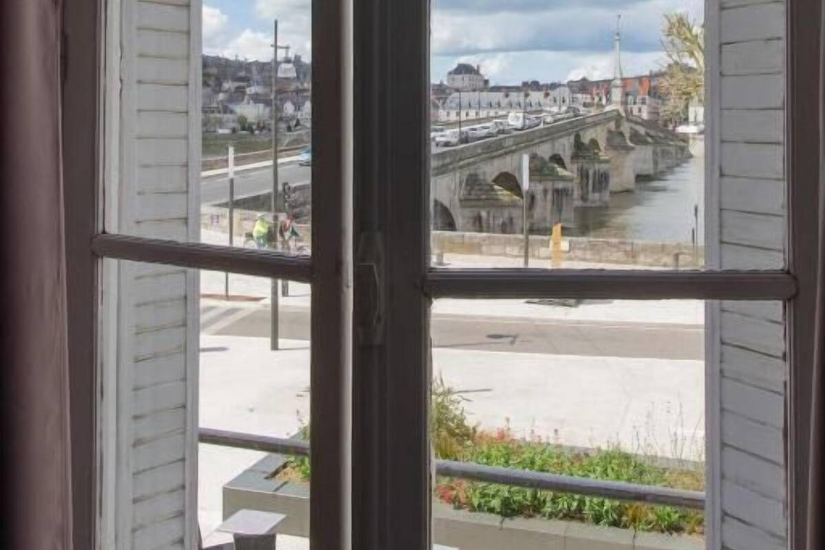 Blick durch ein Fenster mit weißen Fensterläden, der eine Brücke, einen Fluss und eine Stadtlandschaft mit Gebäuden und Grünflächen zeigt.
