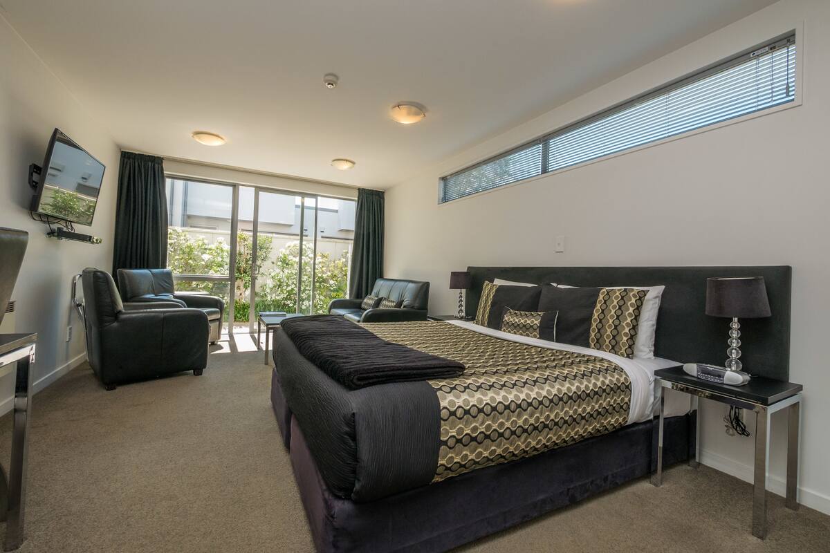 A modern hotel room with a large bed, a sofa, a TV, and a view of the outdoors.