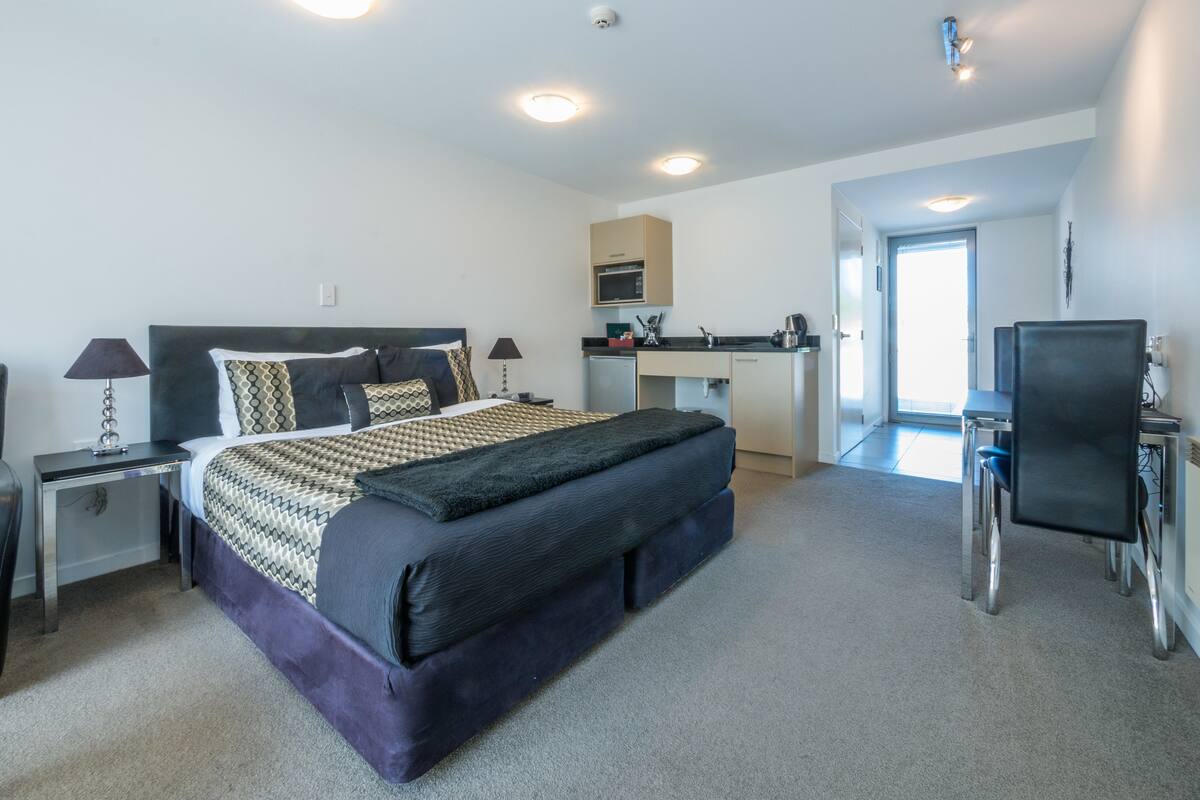 A modern hotel room with a large bed, a desk, and a kitchenette.