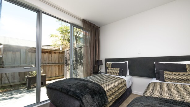 A bedroom with two beds, a large sliding glass door to a backyard, and a view of the outside. - Bellano Motel Suites (Christchurch)