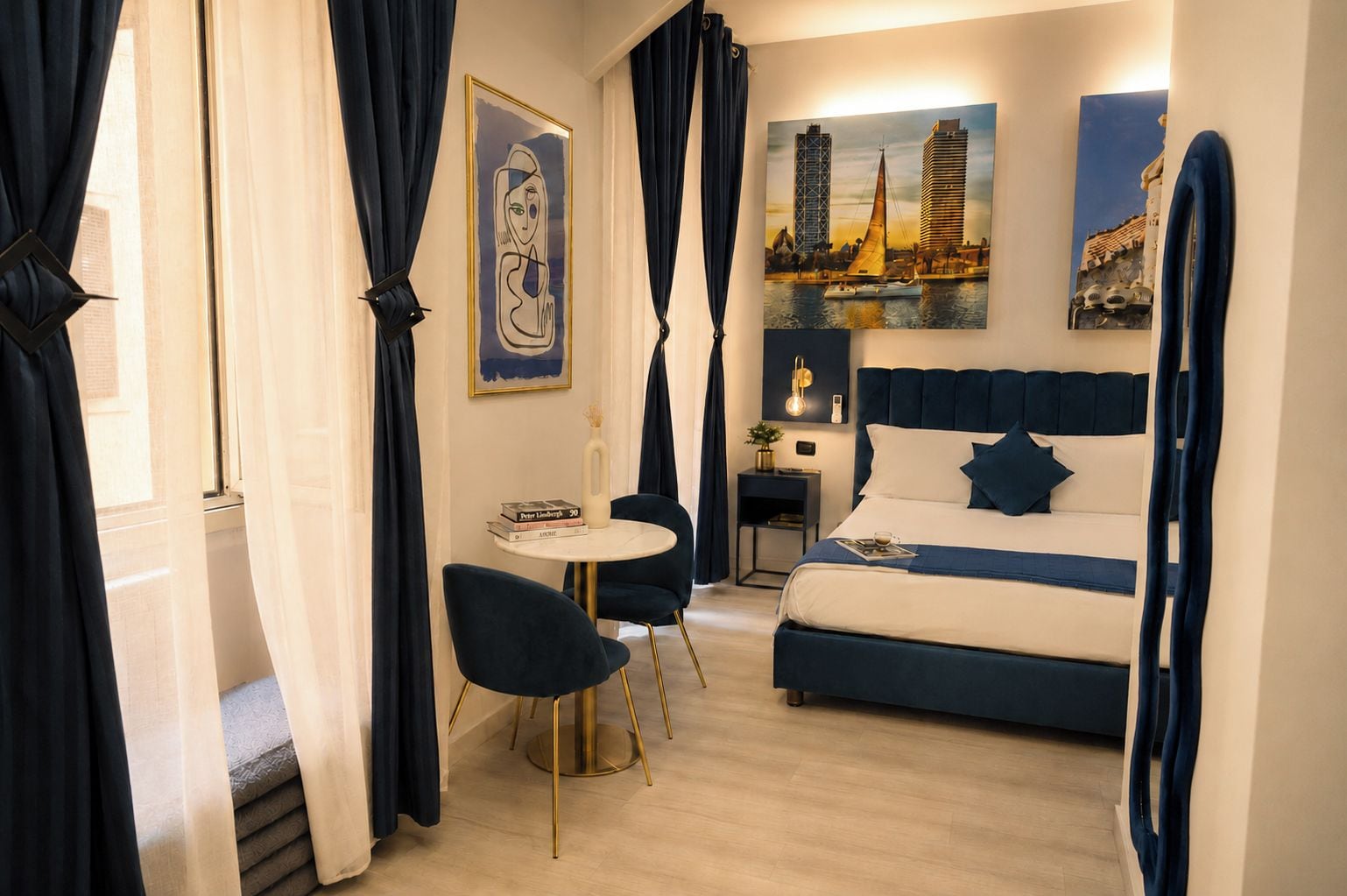 Quarto de hotel com uma cama, uma mesa redonda e uma cadeira. Há cortinas, um quadro e um abajur na parede.