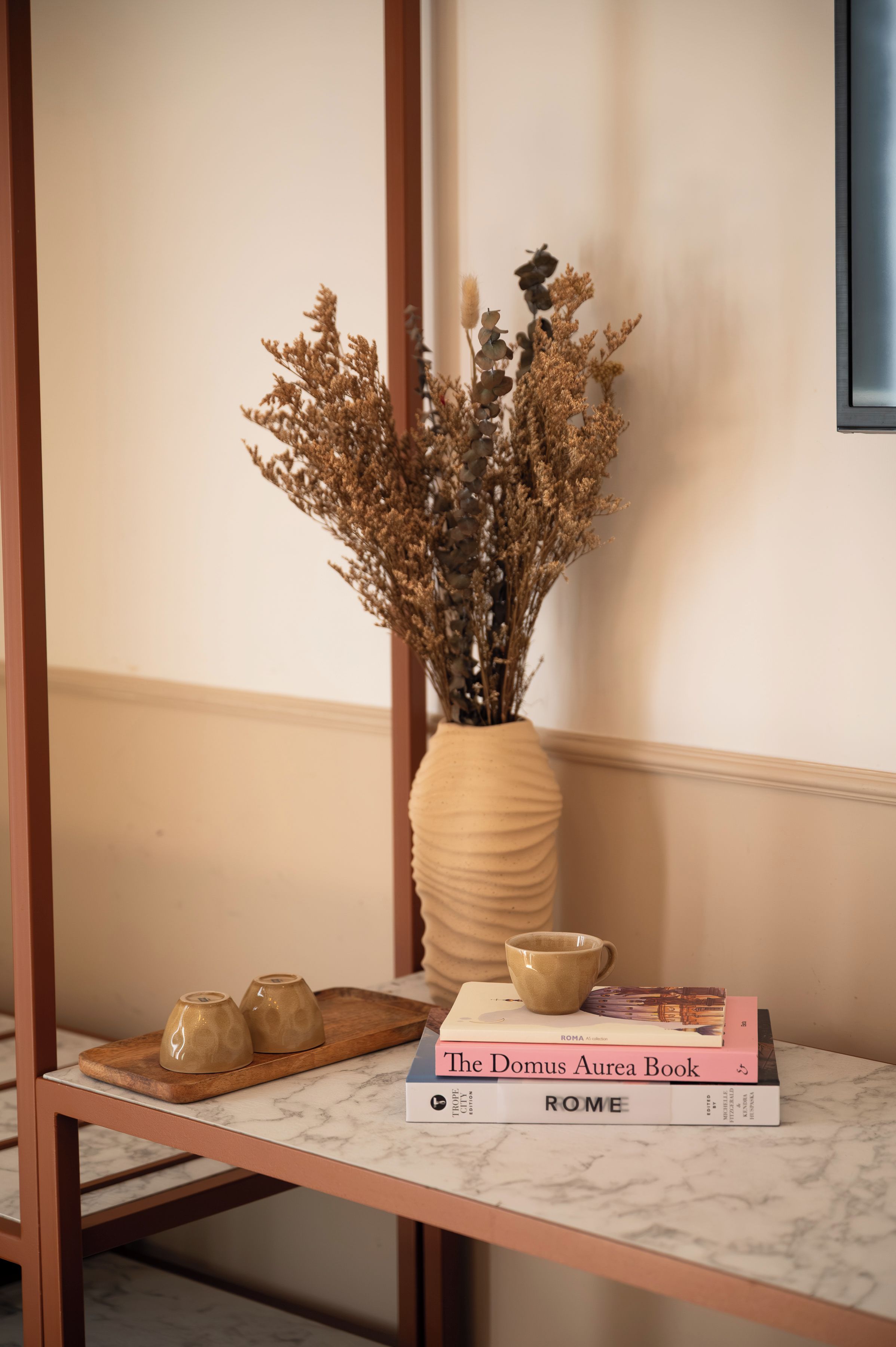 Ein Tisch mit einer Vase getrockneter Blumen, einem Buch mit dem Titel 'The Domus Aurea Book' und zwei Keramiktassen.