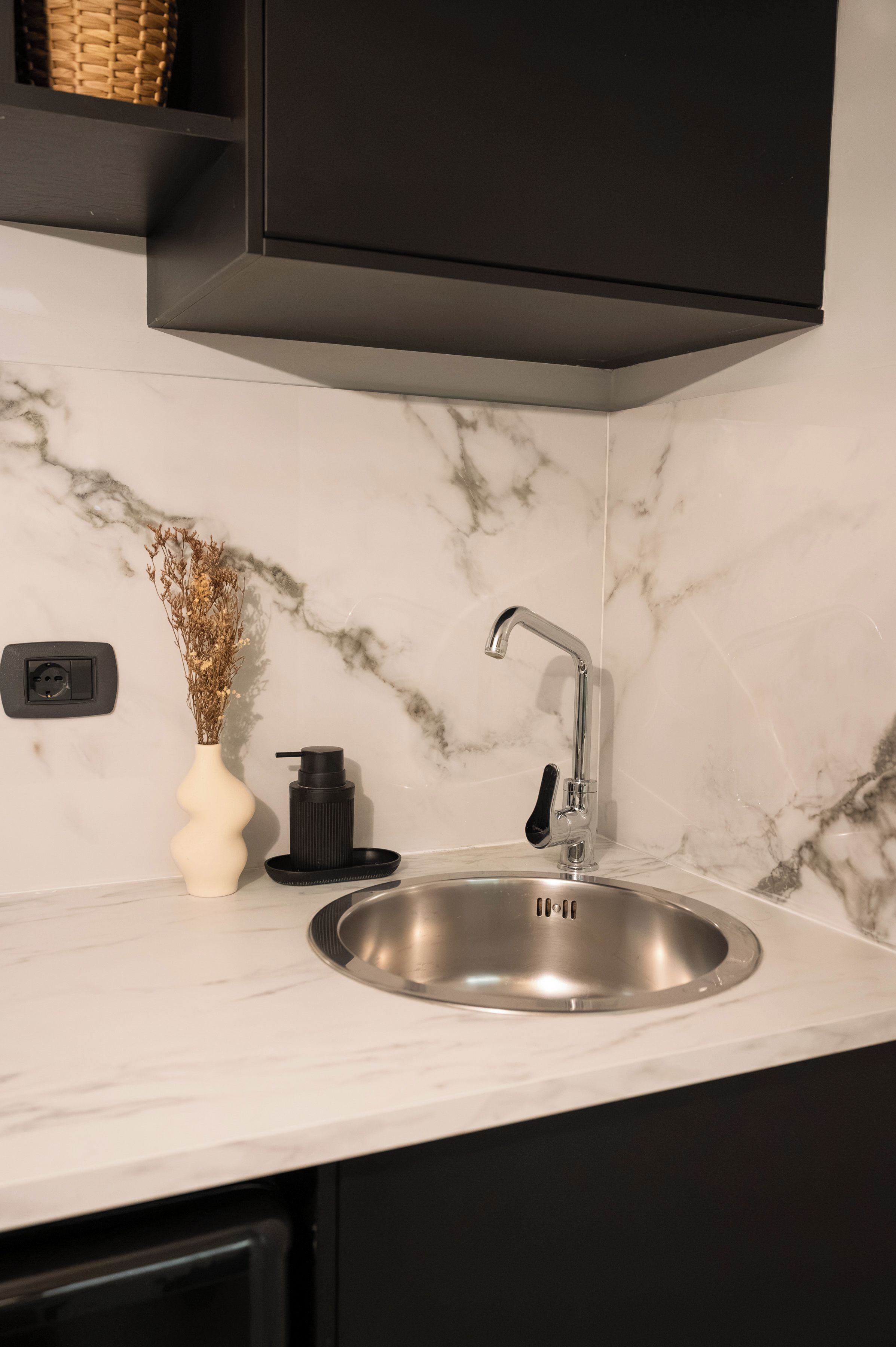 A modern kitchen with a stainless steel sink, a black soap dispenser, and a white vase with dried flowers on the counter.