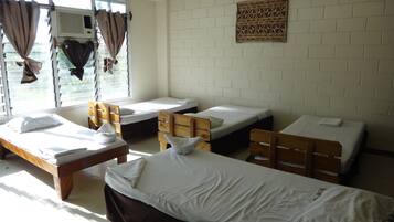 A dormitory room with four single beds, a wooden desk, and a window with curtains.
