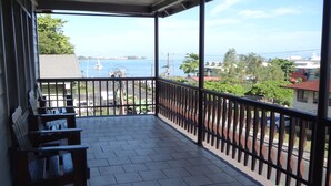 A covered balcony with tiled flooring and a railing, overlooking a body of water and buildings.