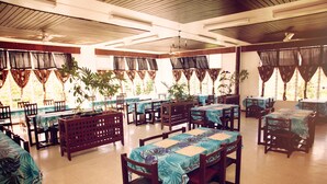 A spacious dining area with multiple tables and chairs, large windows with curtains, and potted plants.