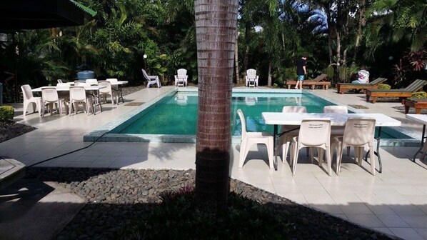 A pool area with white lounge chairs and tables, surrounded by palm trees and greenery.