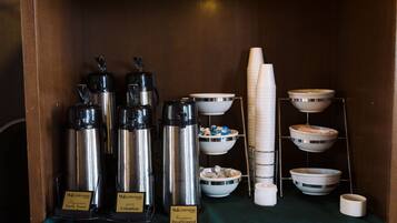 Una estación de café con tres dispensadores de café etiquetados como 'Waldwood Signature Dark Roast', 'Waldwood 100% Colombian' y 'Waldwood Descafeinado'. También hay varios vasos y recipientes blancos desechables con tapas.