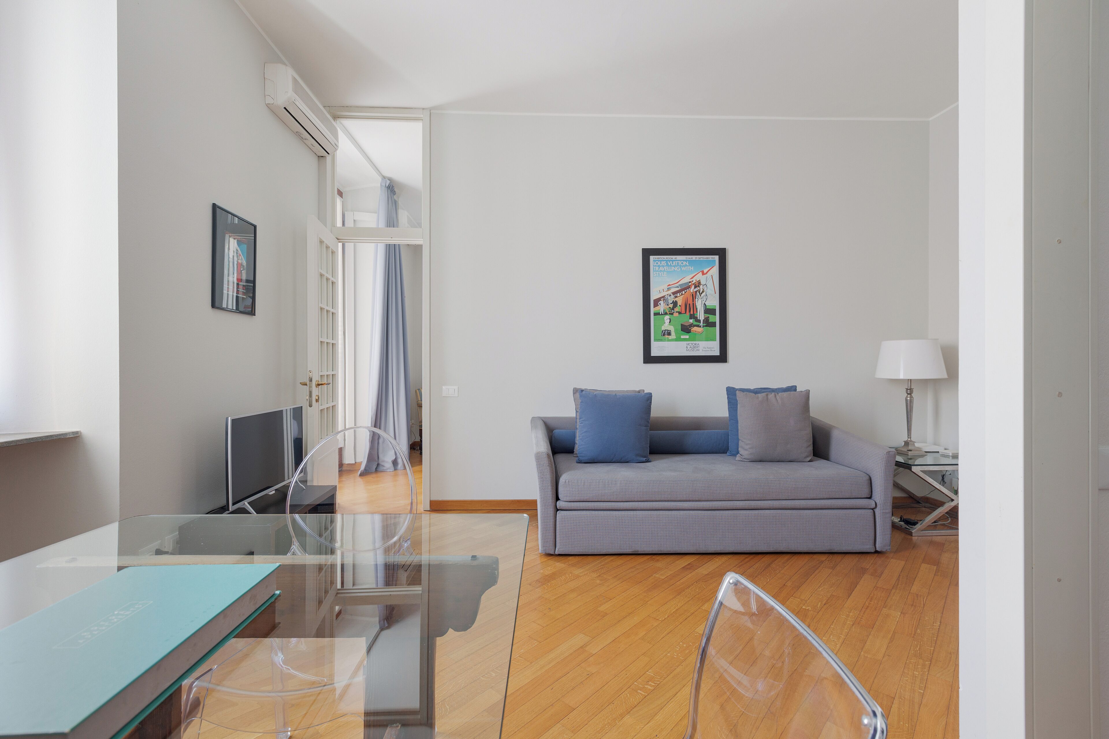 A modern living room with a glass dining table, a grey sofa with blue cushions, and a framed picture on the wall.