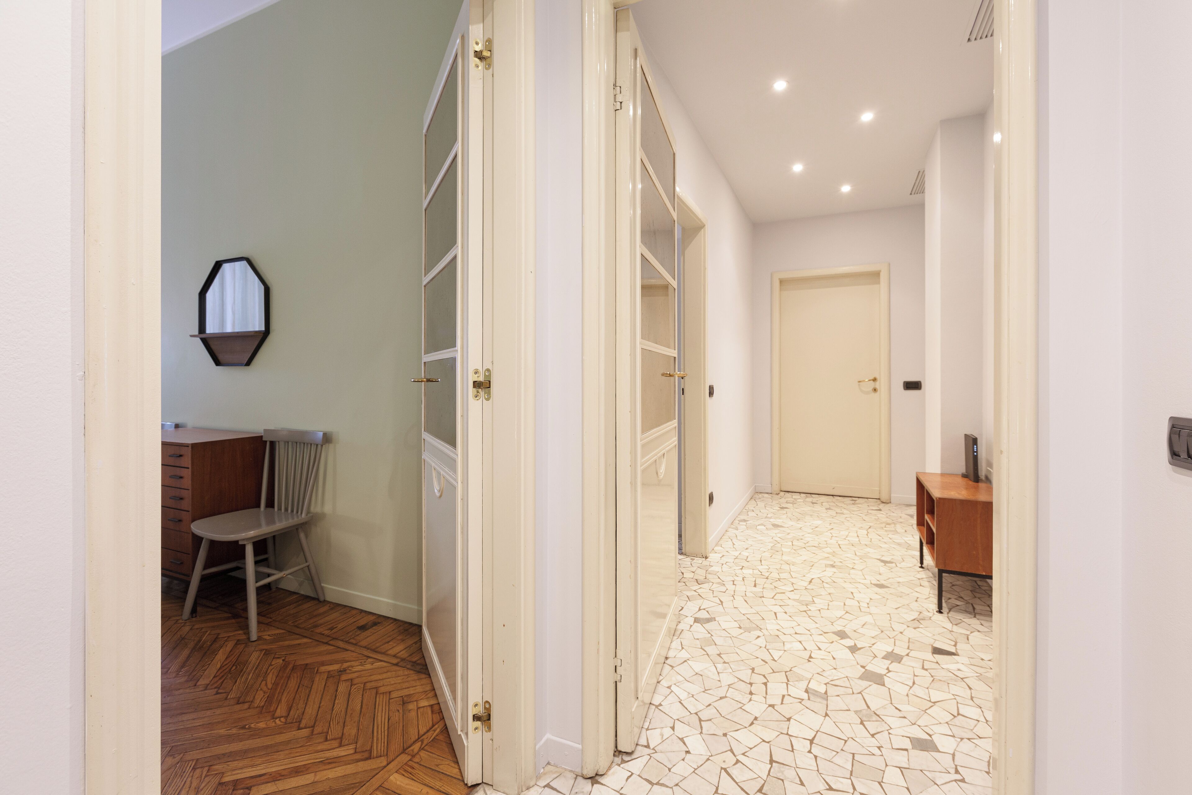 A hallway with a wooden floor, a chair, and a desk with a mirror.