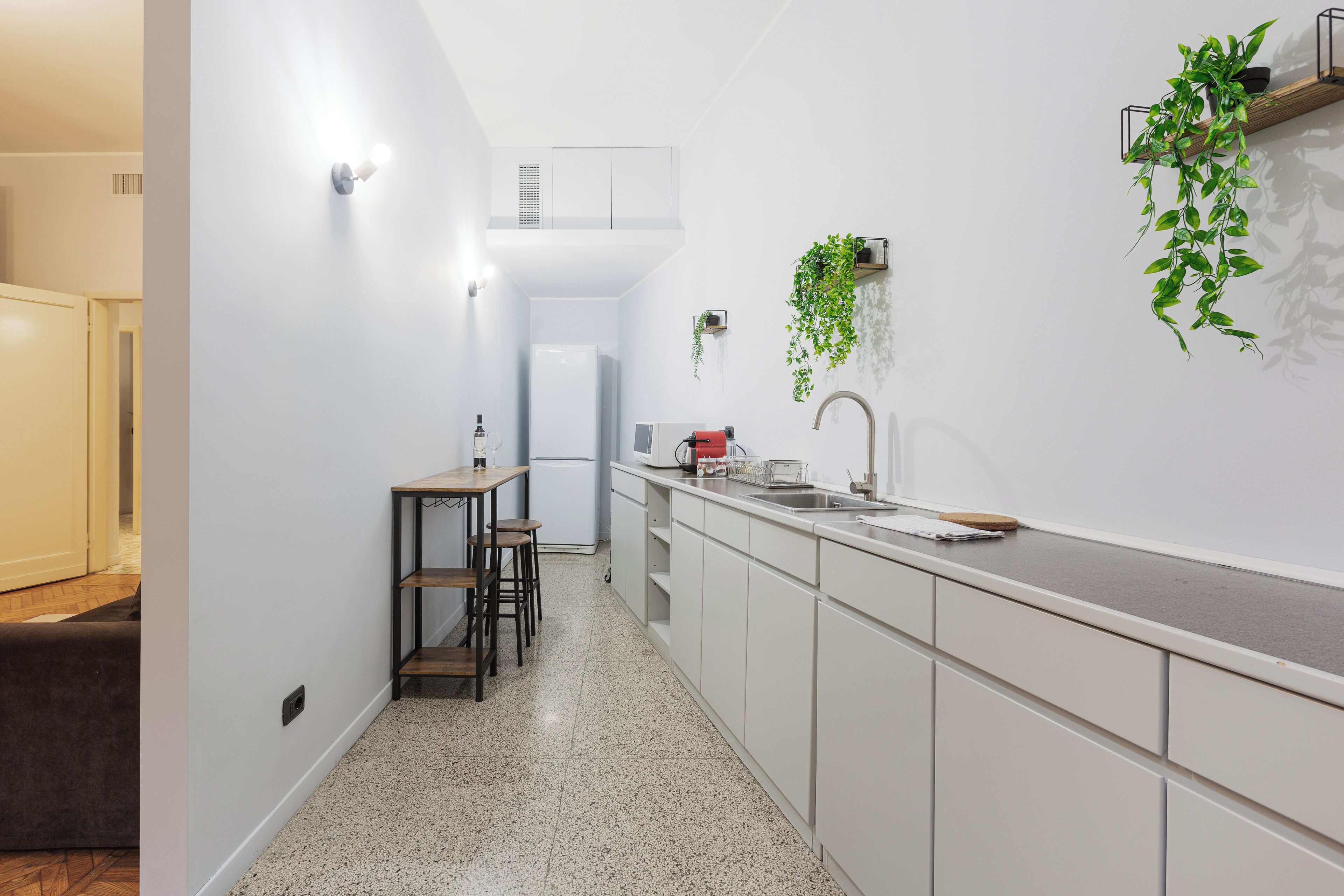 Eine schmale Küche mit weißen Schränken, einer Arbeitsplatte und einem Spülbecken. Zwei Hocker und ein kleiner Tisch sind ebenfalls vorhanden.