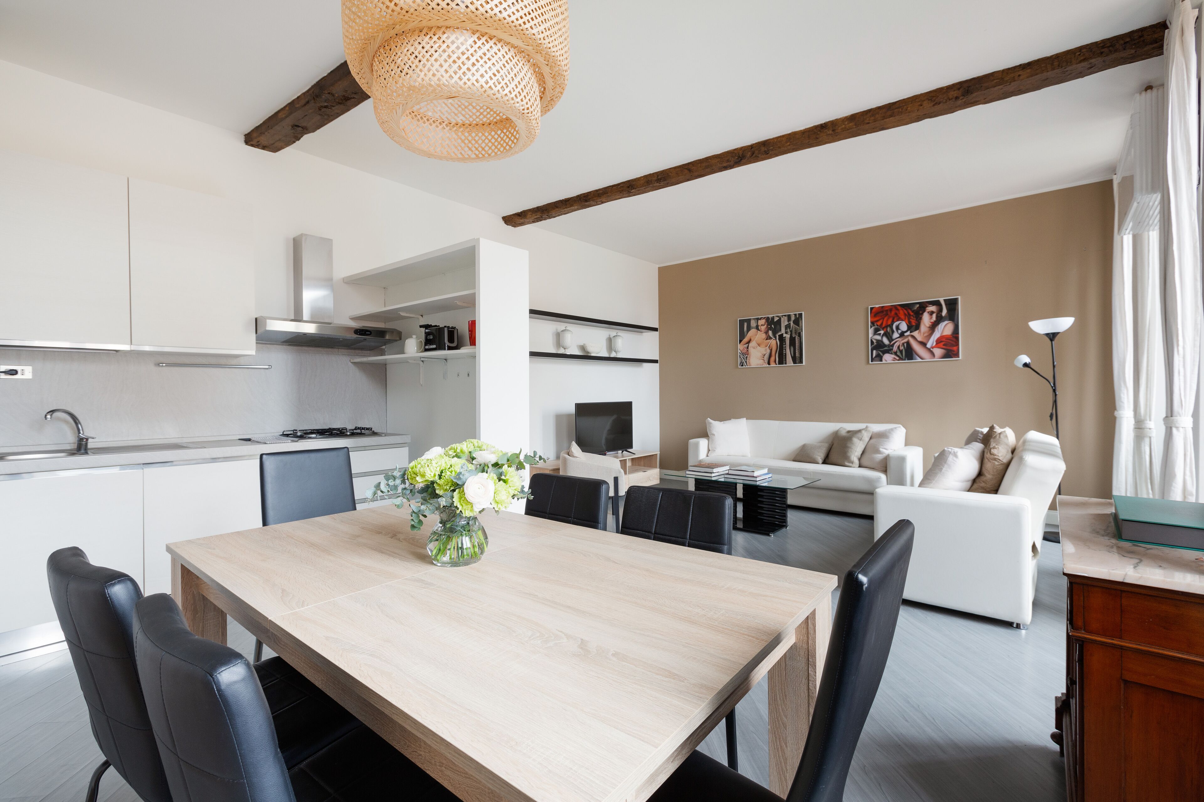 Una cocina moderna con armarios blancos, una mesa de comedor de madera y sillas negras. Hay un sofá blanco y un televisor en la sala.