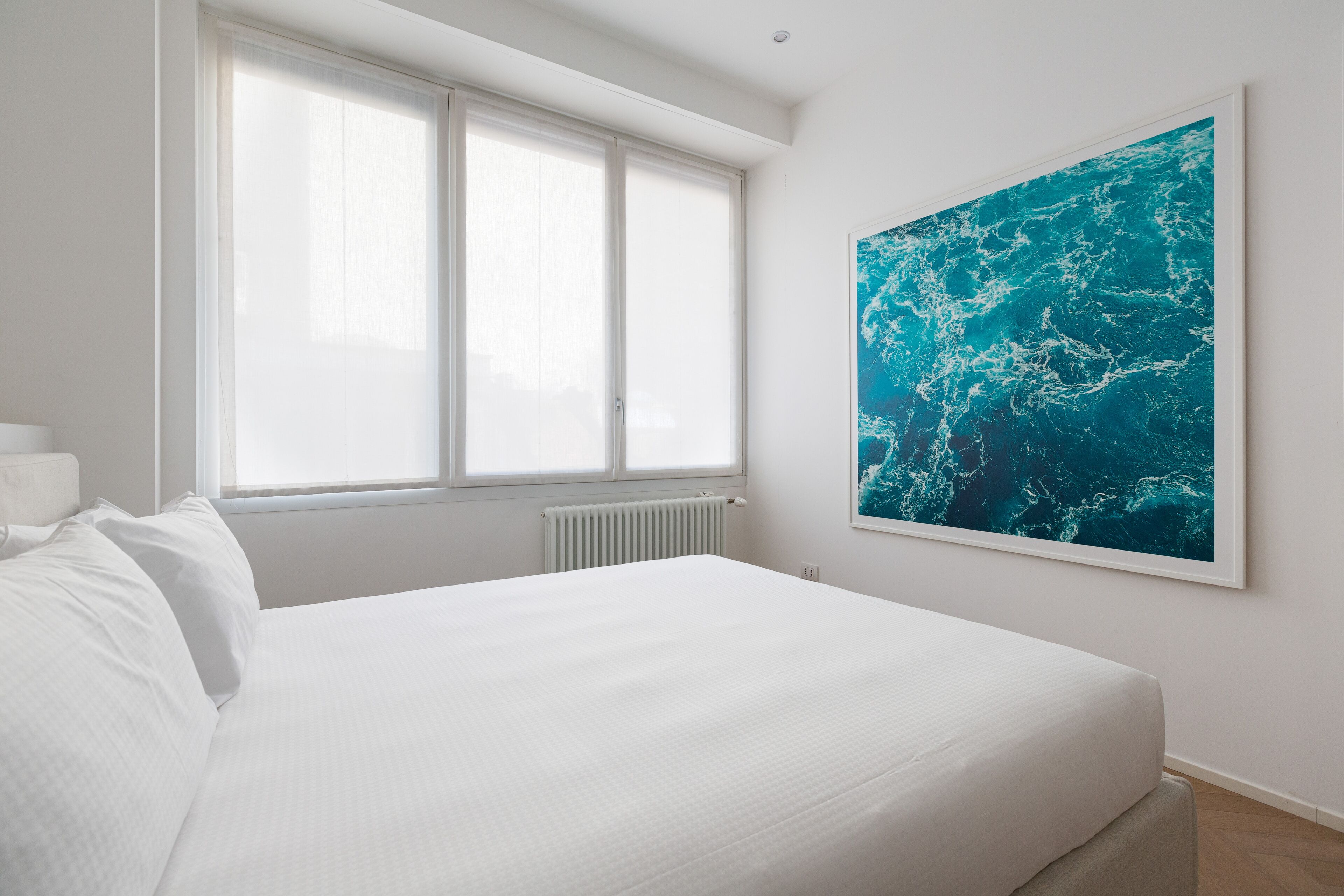 A bedroom with a large bed, a window with blinds, a radiator, and a large ocean-themed painting on the wall.