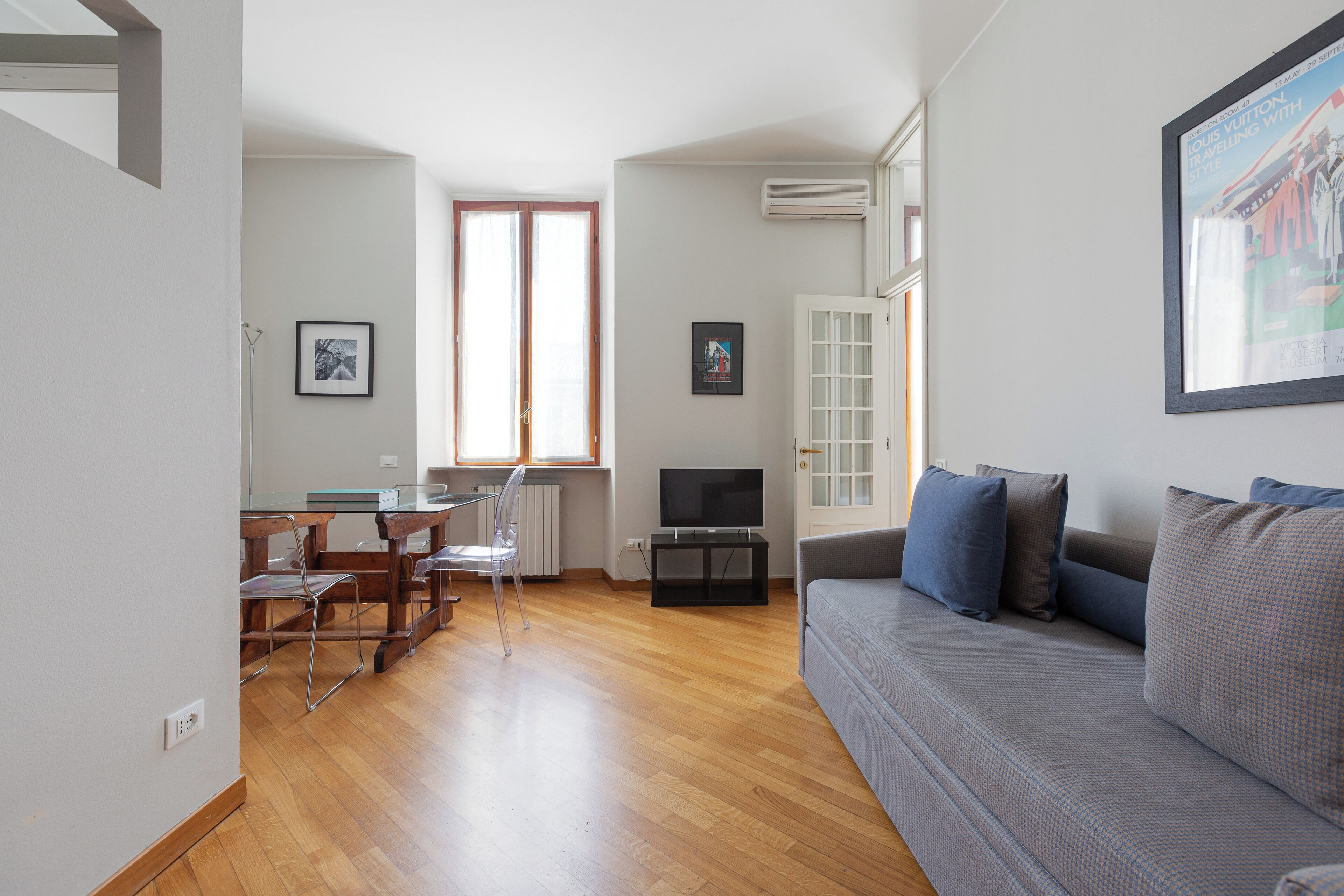 Una sala de estar con un sofá gris, piso de madera, televisor y una zona de comedor con sillas y una mesa.