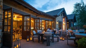 An outdoor patio area with multiple tables and chairs, a bar area visible through large windows, and a stone wall on one side. - Juniper Preserve (Bend)
