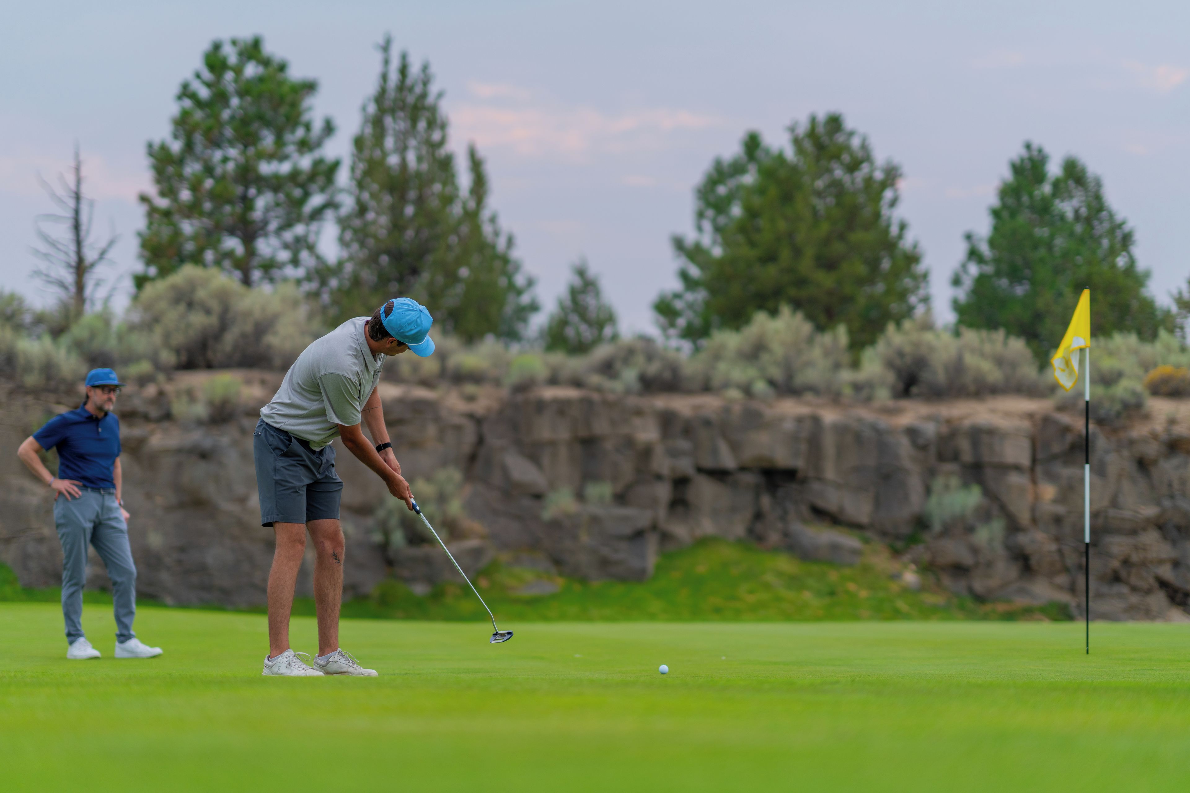 A golf course with a player preparing to putt, another player observing, and a flagstick in the background.