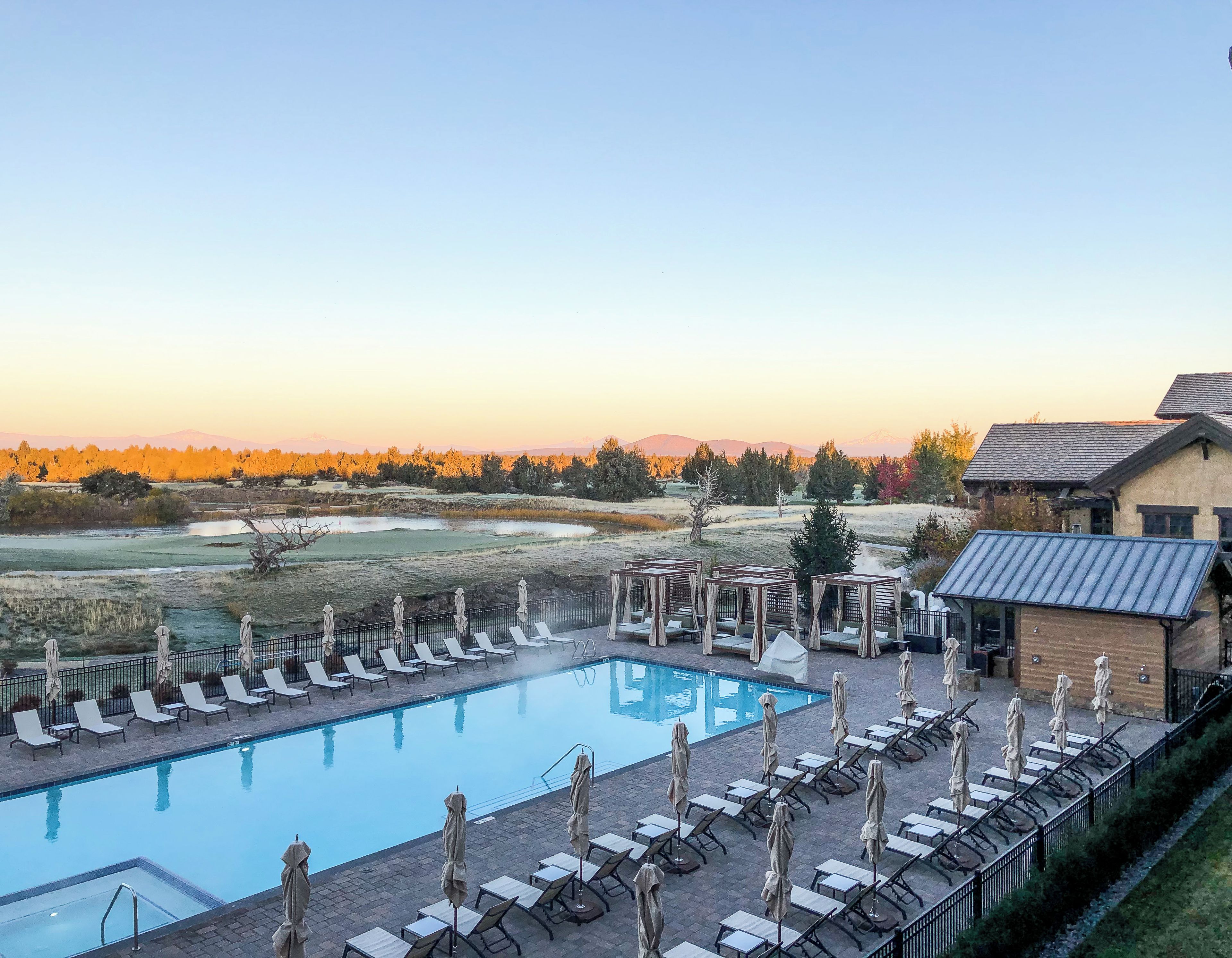 A swimming pool with sun loungers and umbrellas, surrounded by a fence, with a view of trees and distant mountains.