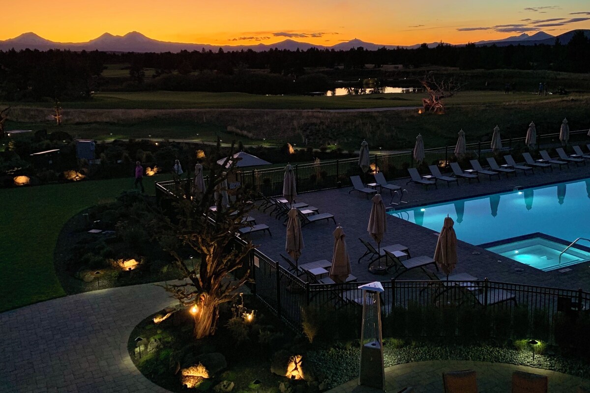Zona de piscina con sillones de descanso y sombrillas, rodeada de jardines ajardinados y con vistas a montañas distantes al atardecer.