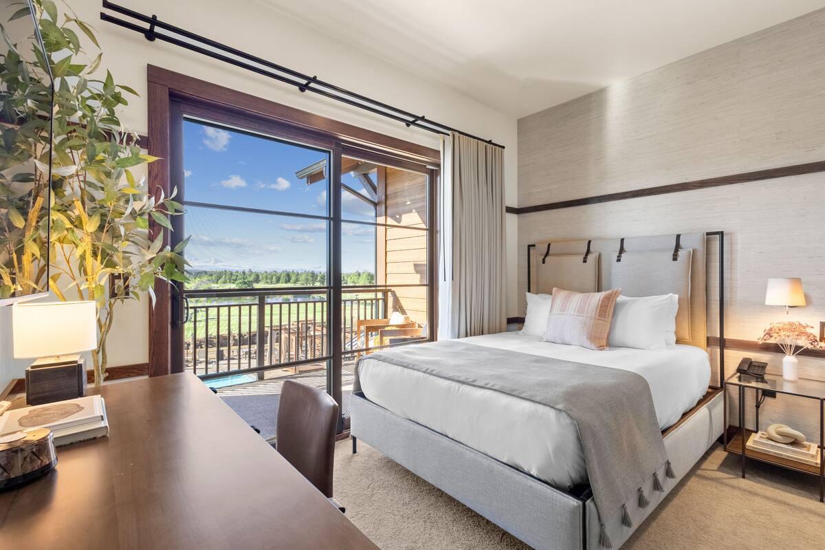 A hotel room with a large bed, a desk with a lamp, a chair, a nightstand with a phone, and a view of the outdoors.