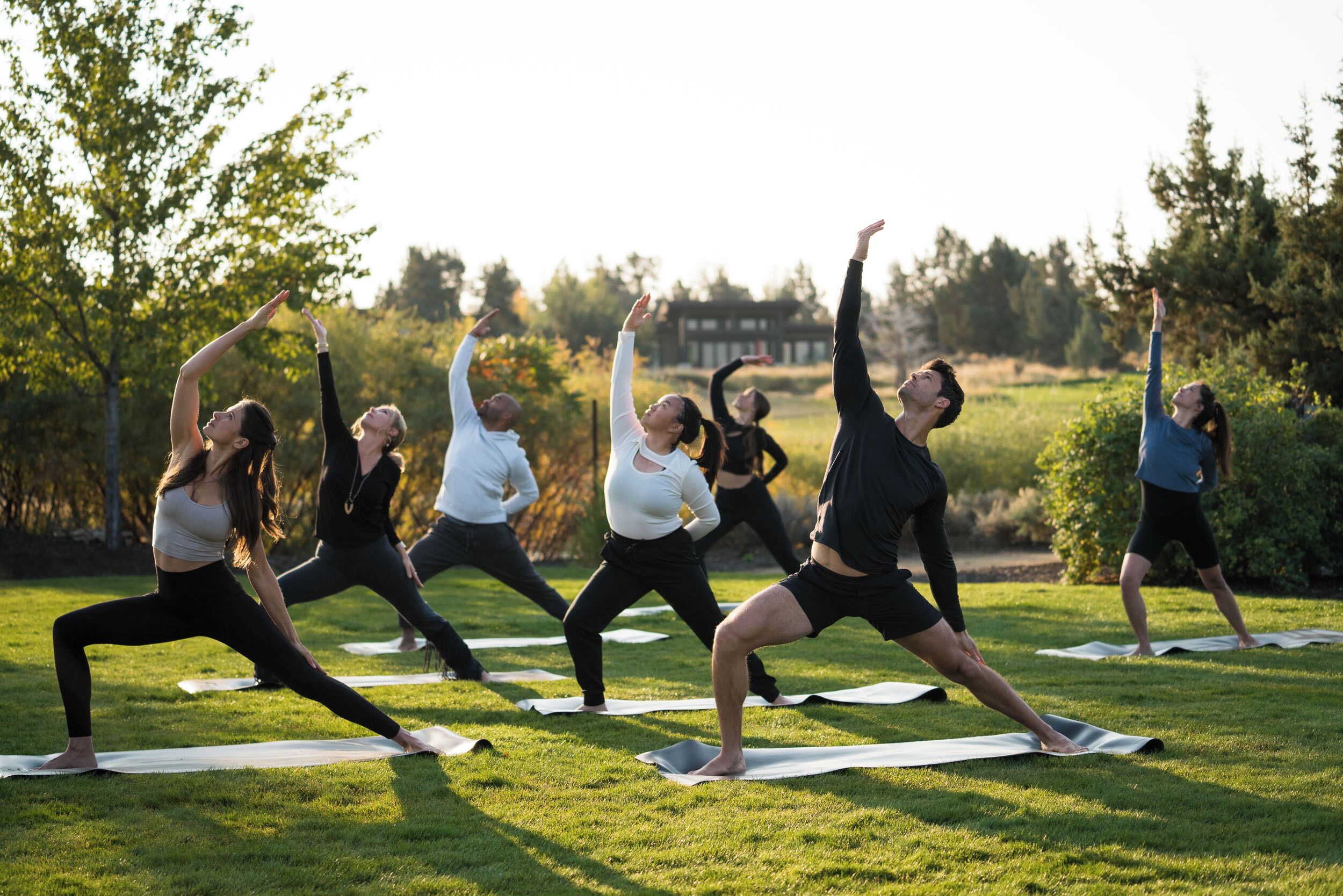 A group of people performing yoga on mats in an outdoor setting with trees and a building in the background.