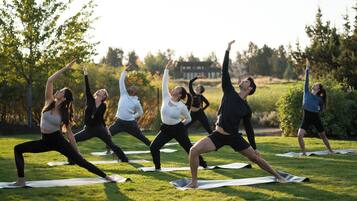 Un groupe de personnes pratiquant le yoga sur des tapis, dans un cadre extérieur, avec des arbres et un bâtiment en arrière-plan.