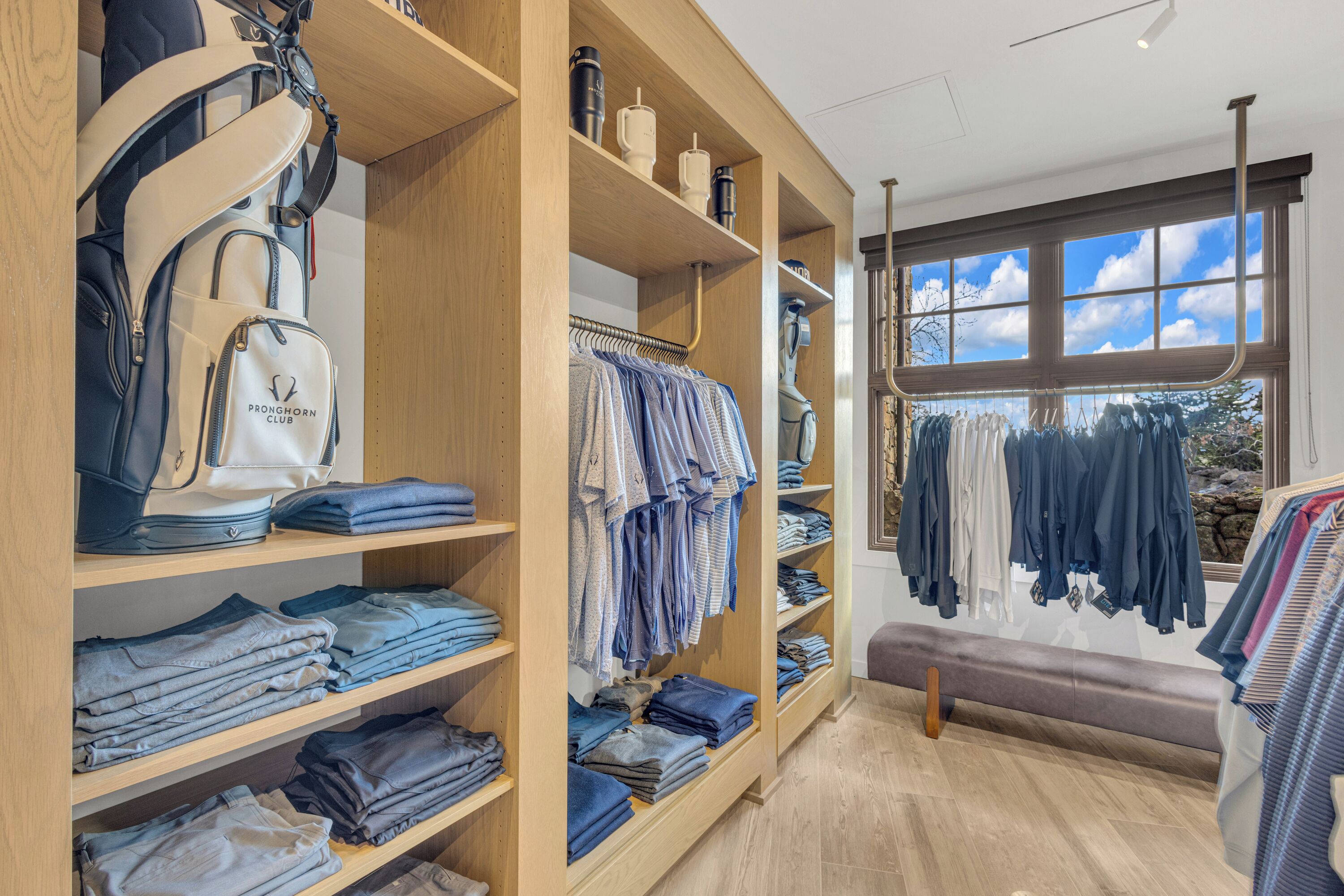 A well-organized clothing store with neatly folded jeans and shirts on shelves, a rack of hanging clothes, and a bench in the center.