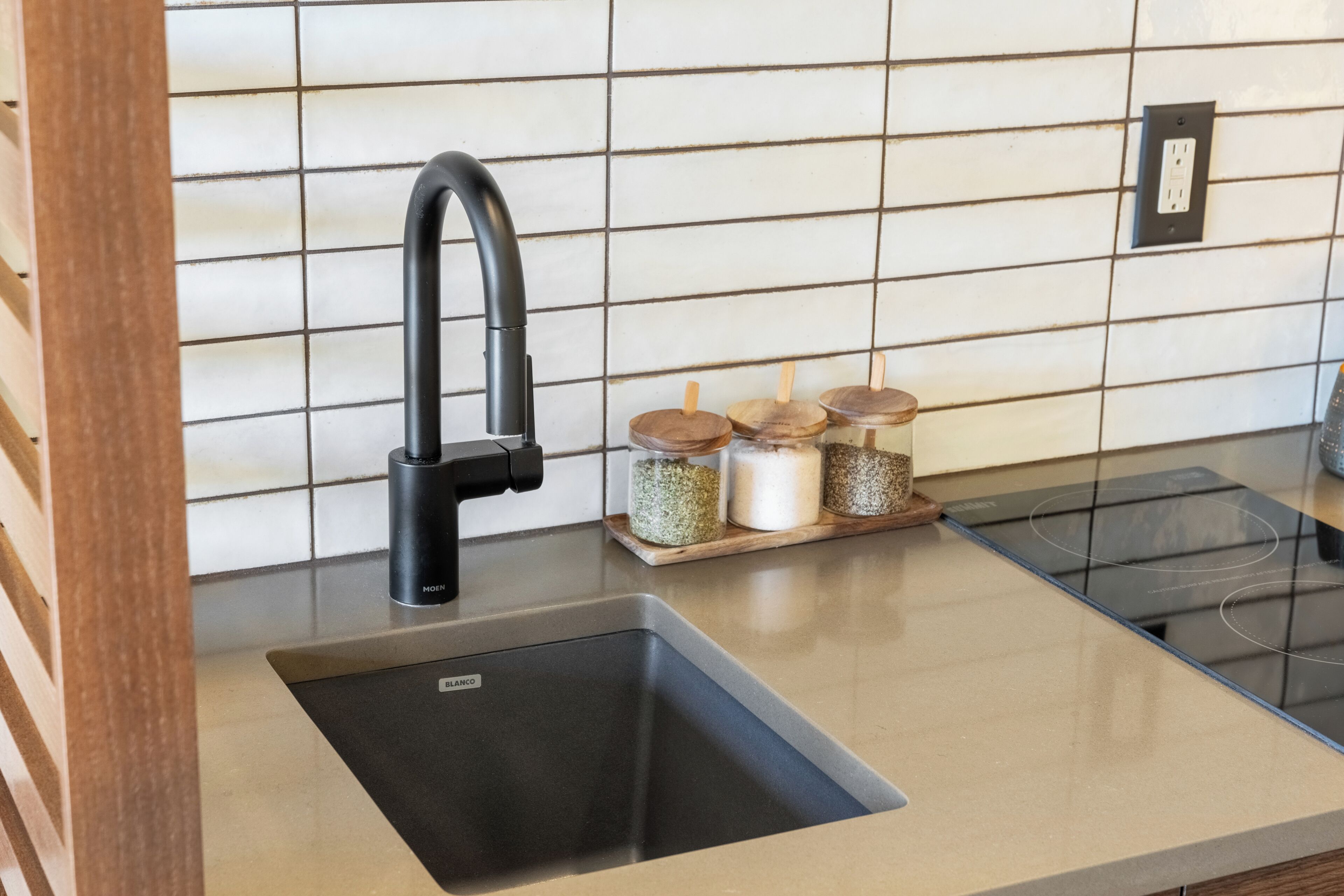 Una cocina con un grifo negro, un lavamanos y frascos de especias de madera sobre una encimera.