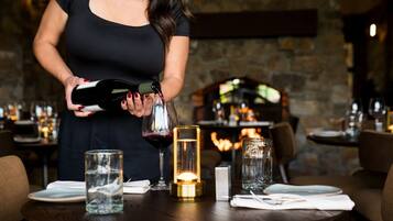 Una persona sirviendo vino en una copa en una mesa de restaurante con velas y chimenea al fondo.